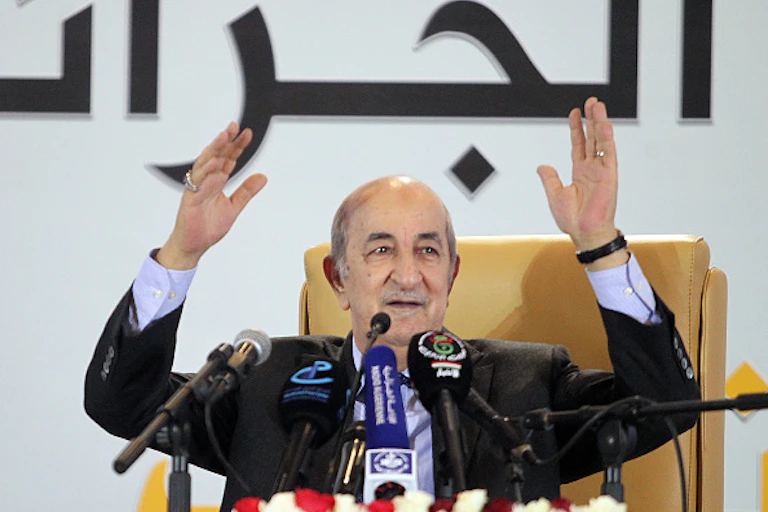 A elderly man sitting behind a table with microphones on it. Both his hands are up.