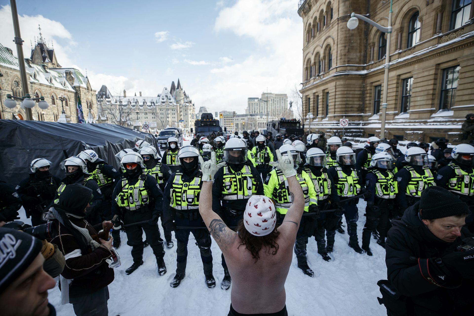 A man naked from the waist up wearing a toque with maple leafs on it confronts a line of police officers.