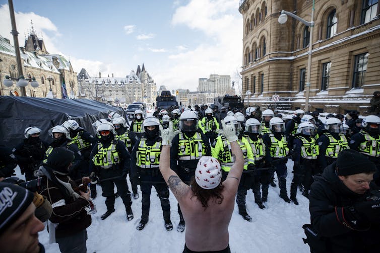 A man naked from the waist up wearing a toque with maple leafs on it confronts a line of police officers.