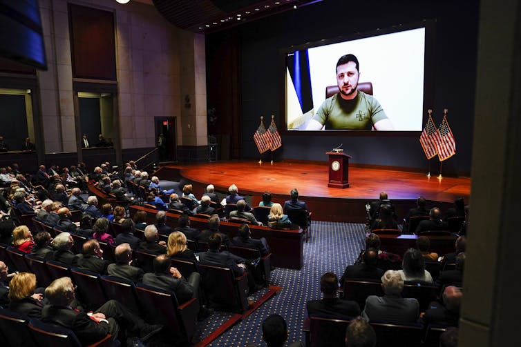 An auditorium full of people look forward at an empty stage and large screen showing Ukrainian President Volodymyr Zelenskyy