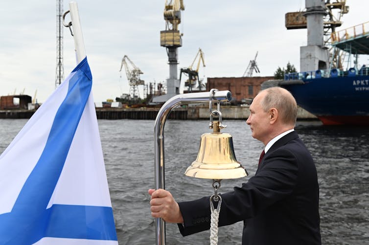 Vladimir Putin stands at the rail of a ship next to a naval flag and a ship's bell.