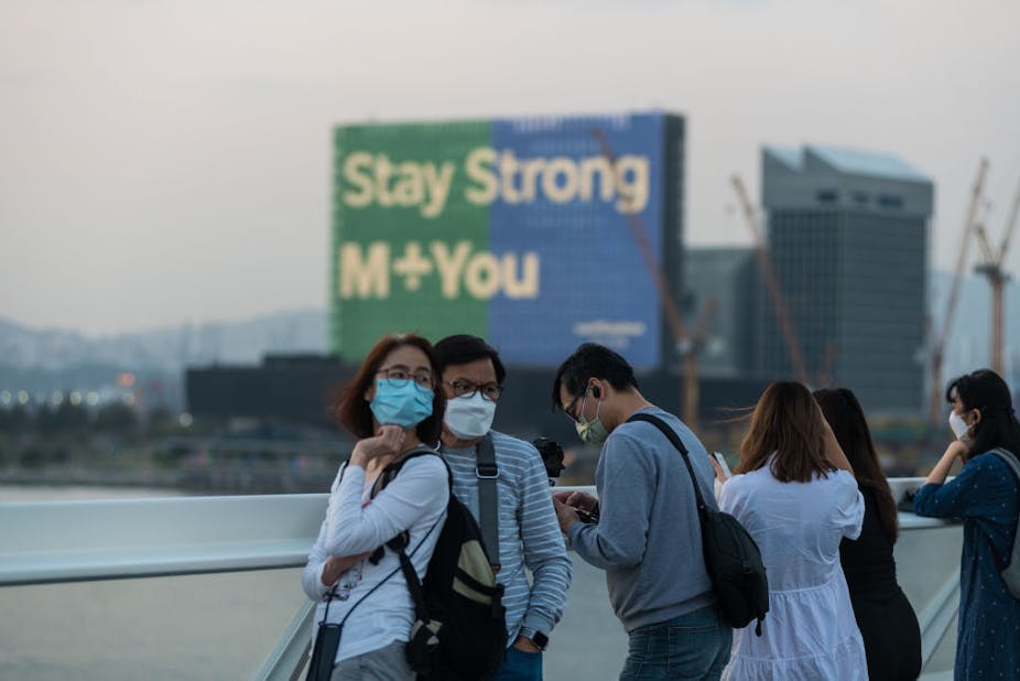 A casually dressed couple wearing surgical masks and glasses pose for a photograph. A billboard behind them reads "Stay Strong M+You"