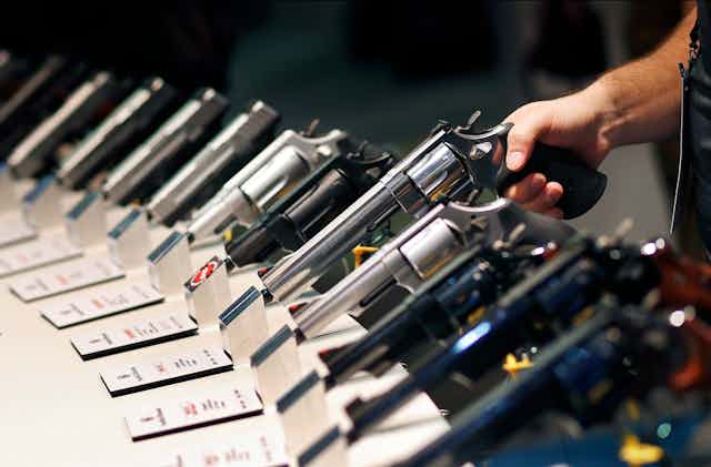A hand lifts a long-barreled handgun from a display rack holding a dozen models.