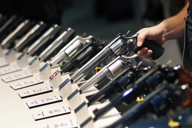 A hand lifts a long-barreled handgun from a display rack holding a dozen models.