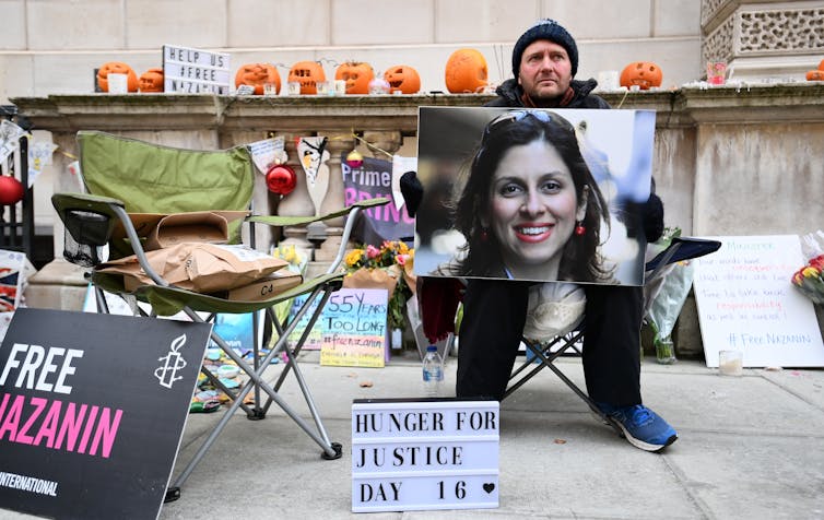 Richard Ratcliffe wears a hat and holds a photo of Nazanin Zaghari-Ratcliffe while sitting in a folding chair. He is surrounded by other photos and signs reading Free Nazanin.