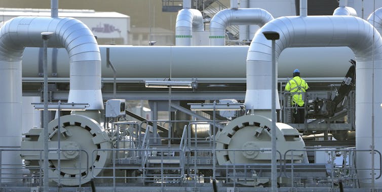 A worker in a yellow fluorescent coat stands next to large pipes that carry gas from Russia to Germany