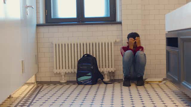Un niño acurrucado en junto a un radiador y una mochila en el cuarto de baño de un colegio.