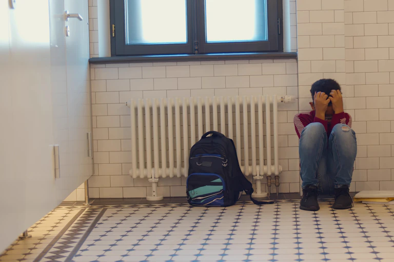 Un niño acurrucado en junto a un radiador y una mochila en el cuarto de baño de un colegio.