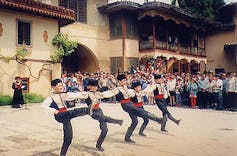 Five boys doing a traditional dance, while locking their arms together, as people watch on the side.