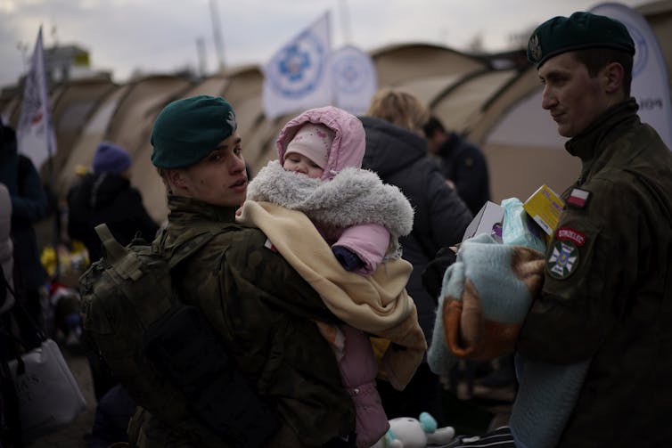 A soldier holds a bundled up baby while another soldier stands beside him with a bag