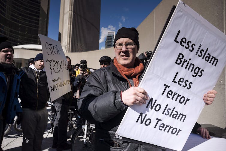 A man in a black cap and wearing glasses carries an anti-Muslim sign.