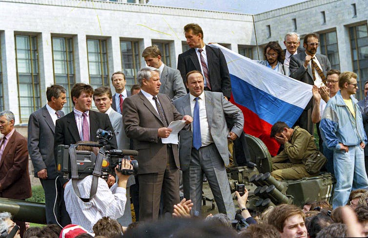 A man in a suit standing on top of a tank and holding a small stack of paper. He is surrounded by several other men in suits and there is a news camera pointed at him. Behind him is the Russian flag.