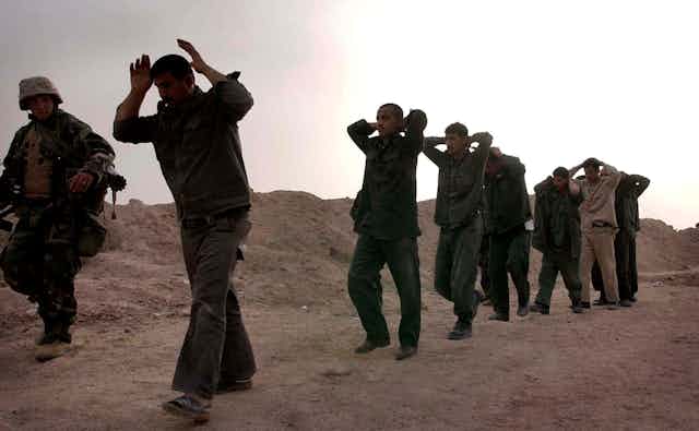 Iraqi soldiers walk with their hands over their heads next to a U.S. Marine along a dusty path.