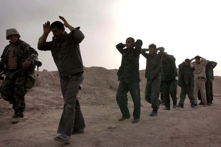 Iraqi soldiers walk with their hands over their heads next to a U.S. Marine along a dusty path.