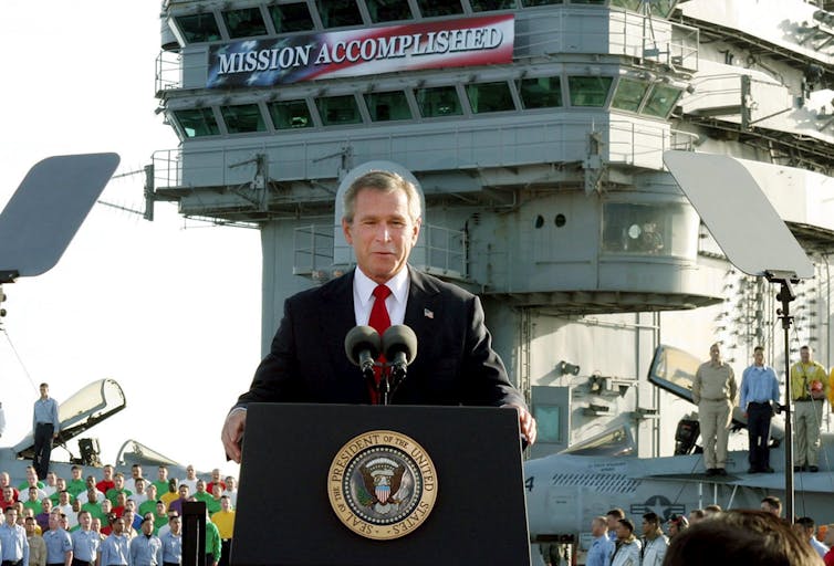 A grey-haired man speaks at a podium with a banner over his head reading mission accomplished