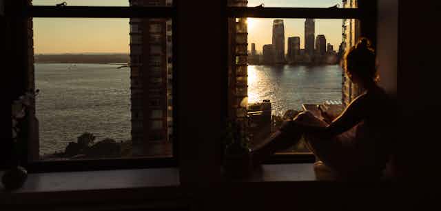 Woman in a dark room sits near a window looking at a body of water, buildings in the distance and a low sun that could be either rising or setting.