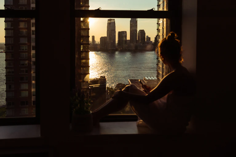 Woman in a dark room sits near a window looking at a body of water, buildings in the distance and a low sun that could be either rising or setting.