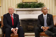 A man with orange-blond hair in a dark suit and red tie smiles sitting next to a Black man in a dark suit and patterned blue tie.