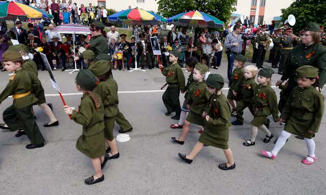 Children wearing army-green uniforms march past veterans in a parade.