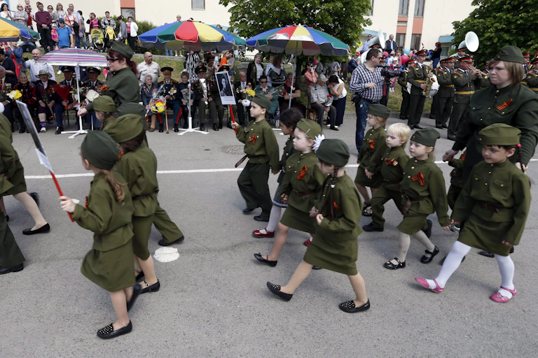 Children wearing army-green uniforms march past veterans in a parade.