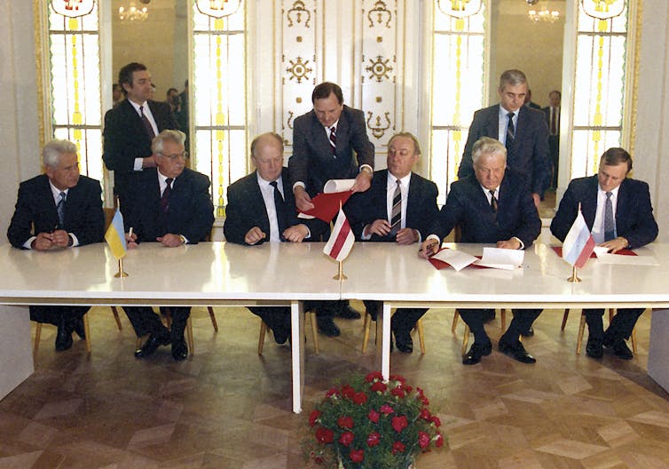 Six men in suits sit behind a table. One of the men is signing a document while another is being handed a similar document. The Russian, Belarussian and Ukrainian flags sit on the table in front of the men.