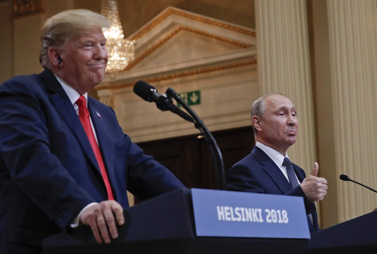 a man with blond-grey hair smiles as a bald man next him gives a thumb's up. both stand in front of podiums that say helsinki 2018.