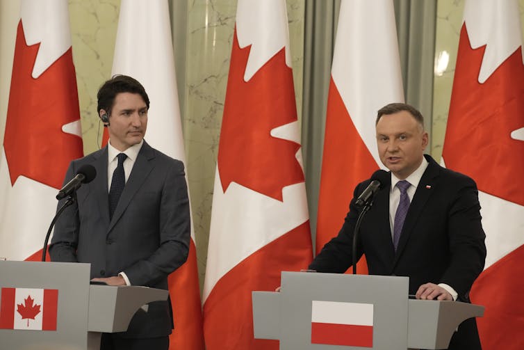 Two men stand behind separate podiums with a row of Canadian and Polish flags behind them.
