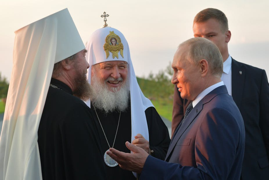 Two men wearing Orthodox Christian headdresses talk with Vladimir Putin.