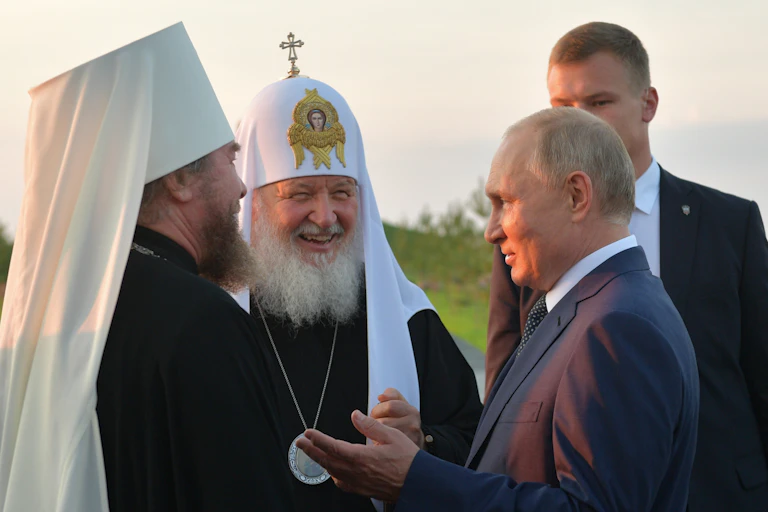 Two men wearing Orthodox Christian headdresses talk with Vladimir Putin.