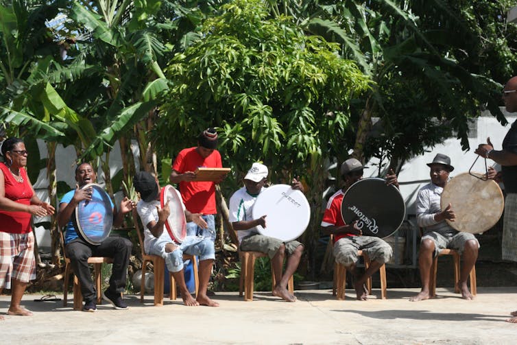 Musicians sit on chairs outdoors playing and singing.