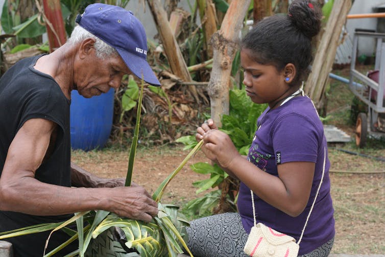 A girl in a purple top watches as a old man with white hair and a blue cap weaves a basket.