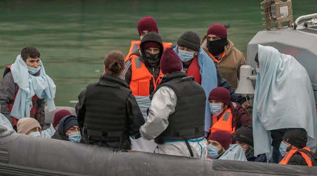 A group of migrants, mostly men, on a Border Force vessel. The people are wearing woolly hats and life jackets, others are wrapped in blankets
