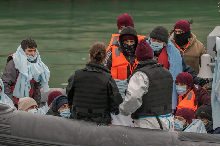 A group of migrants, mostly men, on a Border Force vessel. The people are wearing woolly hats and life jackets, others are wrapped in blankets