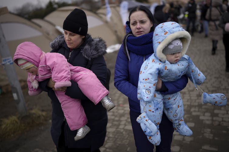 Two women carry a baby each, one in a bright pink snowsuit and the other in a pale blue patterended snow suit.