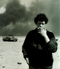 A young man smokes a cigarette in the desaert with a burned tank and smoke in the background.