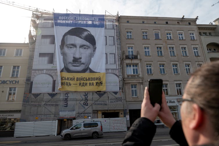 A man taking a picture of a large banner hanging from a building that shows Vladimir Putin's face mocked up to look like Adolf Hitler.
