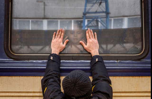 A man presses his palms against a train window as his daughter looks down at him through the glass.