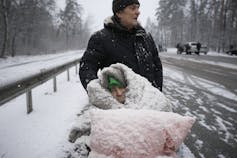 An elderly woman is coated in snow as she sits in a wheelchair under a pink blanket.