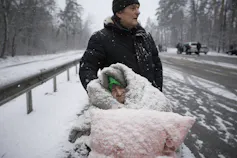 An elderly woman is coated in snow as she sits in a wheelchair under a pink blanket.