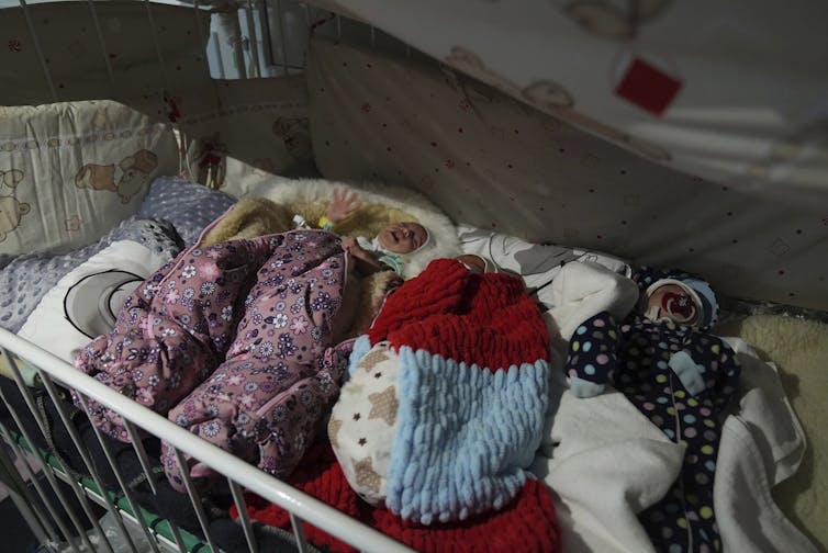 A row of babies swaddled in blankets in a crib, one crying.