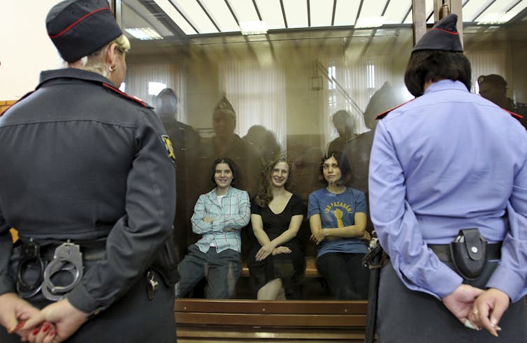 Three women behind a glass panel look out at a courtroom.