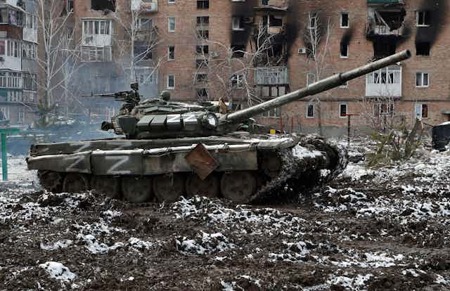 A Russian tank marked with the letter Z sits in the courtyard in front of a bombed apartment building