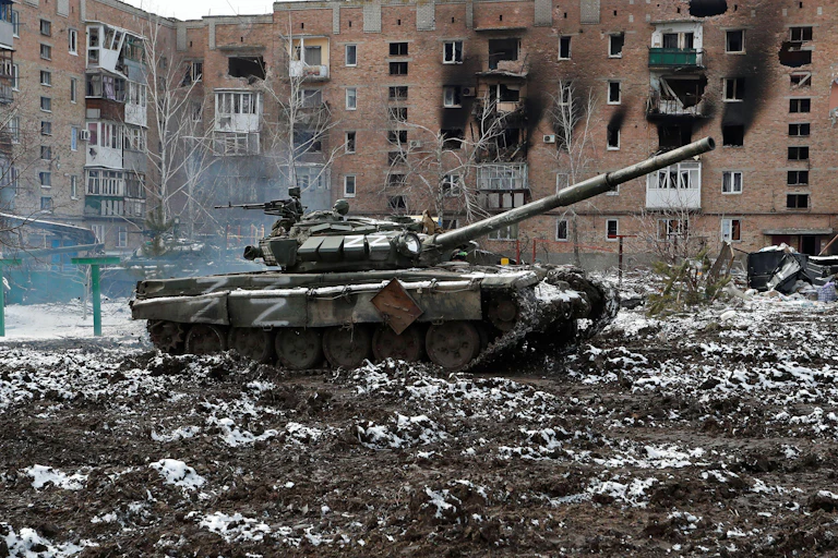 A Russian tank marked with the letter Z sits in the courtyard in front of a bombed apartment building