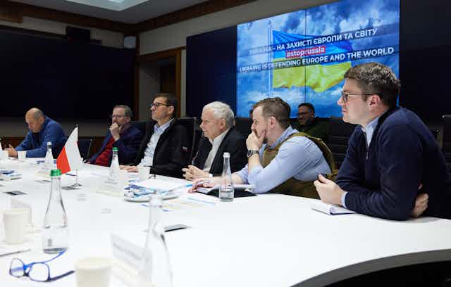 Six white men who are leaders of European countries are seen talking at a table underneath a television screen displaying the Ukrainian flag.