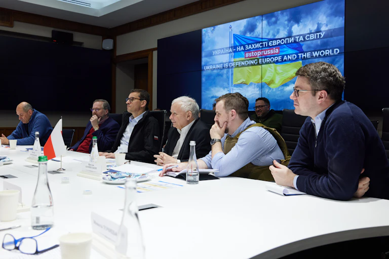 Six white men who are leaders of European countries are seen talking at a table underneath a television screen displaying the Ukrainian flag.