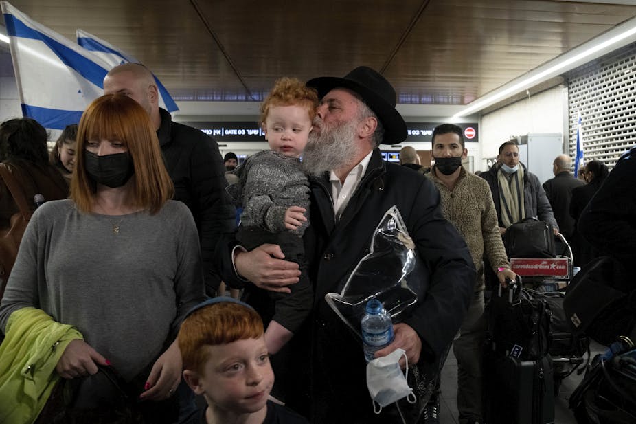 A rabbi with a beard holding a child in his arms with a woman and another child next to him and several people walking behind him.