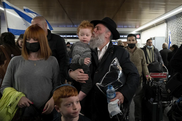 A rabbi with a beard holding a child in his arms with a woman and another child next to him and several people walking behind him.