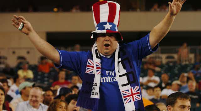 A Chelsea fan wearing a scarf and a 'stars and stripes' hat holds his arms aloft.