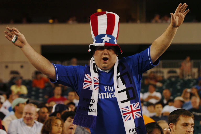 A Chelsea fan wearing a scarf and a 'stars and stripes' hat holds his arms aloft.