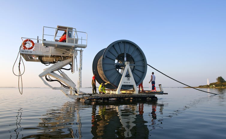 A crew works on a machine with a giant spool laying a cable in the water